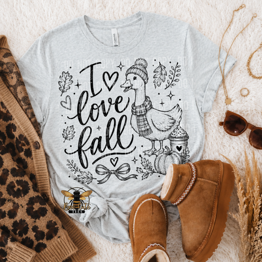 A light heather gray graphic T-shirt laid flat on a white textured rug with fall-themed styling props, including a brown leopard-print sweater, tan suede boots, sunglasses, and dried grass. The shirt graphic reads “I love fall” with a duck wearing a beanie and scarf, plus small autumn motifs.