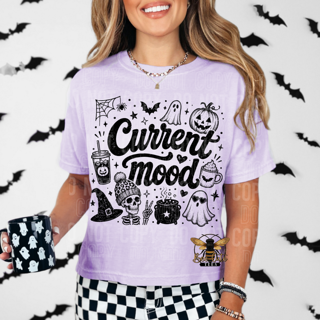 A woman wearing a light purple short-sleeve graphic T-shirt with black Halloween-themed illustrations and the words 'Current mood' across the front. She is holding a black mug with a white ghost pattern, set against a white wall decorated with black bat silhouettes.