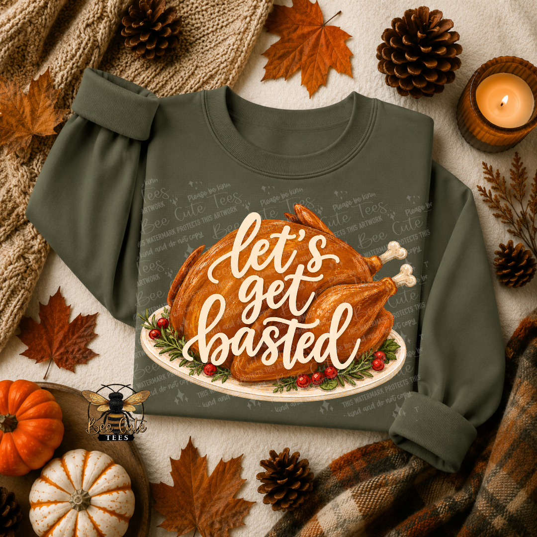 An olive green crewneck sweatshirt with a graphic of a roasted turkey on a platter and the text 'let's get basted' in white script. The sweatshirt is styled in a cozy autumn flat-lay setting with pumpkins, pinecones, fall leaves, a knitted blanket, and a lit candle.