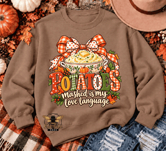 A brown crewneck sweatshirt with a colorful autumn-themed potato graphic, styled in a flat lay with pumpkins, a hat, and denim.