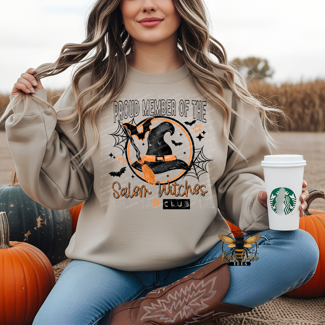 A woman sitting outdoors among pumpkins wears a beige sweatshirt with a Halloween graphic that reads 'Proud Member of the Salem Witches Club.' She holds a white coffee cup.