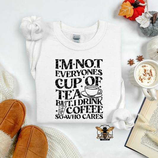 A white long-sleeve t-shirt with a black graphic text print reading 'I'M NOT EVERYONE'S CUP OF TEA BUT I DRINK COFFEE SO WHO CARES' with a coffee cup illustration. The shirt is displayed in a cozy flat-lay arrangement with a mug of coffee, knit socks, slippers, a book, and autumn leaves.