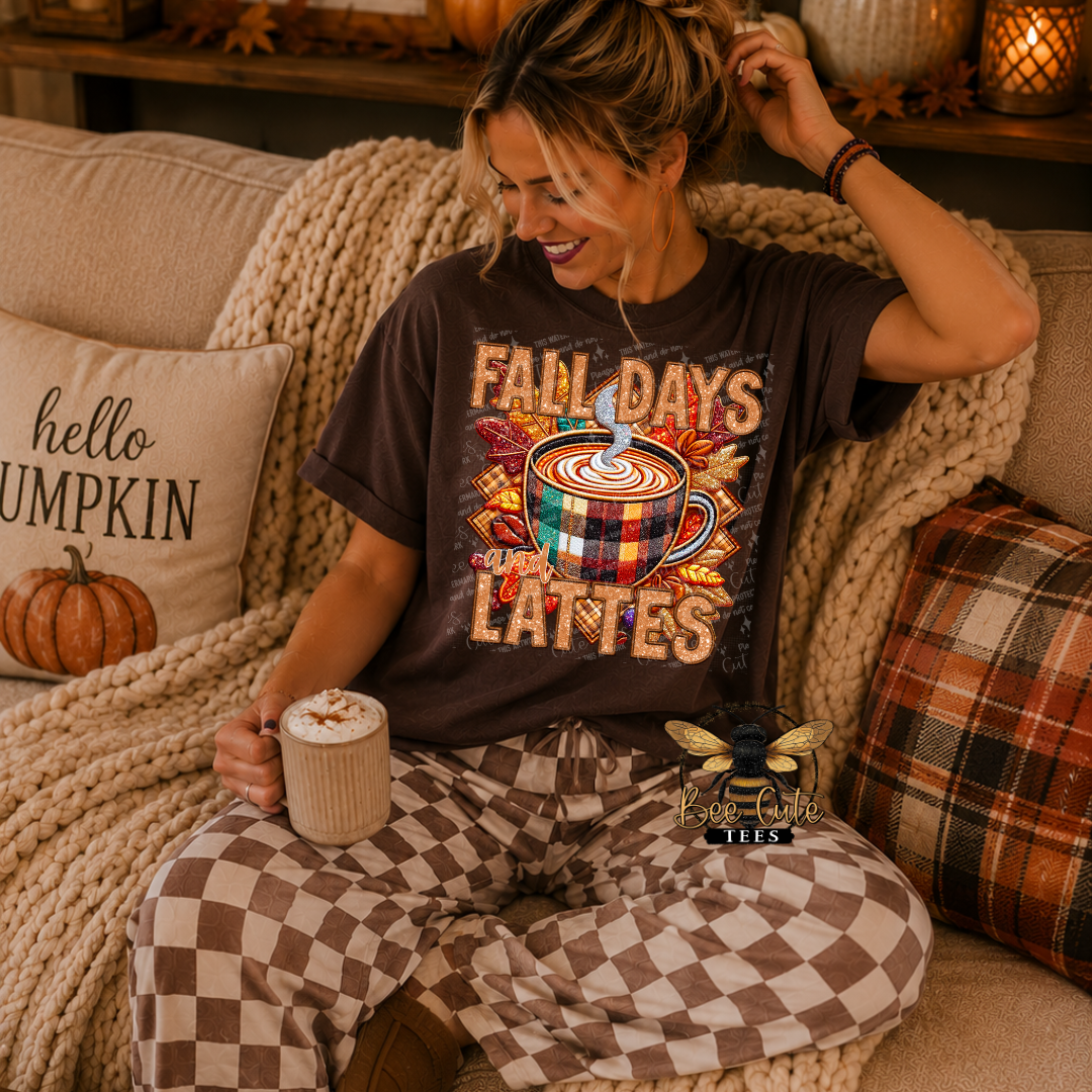 A woman sitting on a cozy couch wearing a dark brown graphic t-shirt that reads 'FALL DAYS LATTES' with a coffee cup design, paired with brown and beige checkered pajama pants. She is holding a mug of coffee, surrounded by autumn-themed pillows and a chunky knit blanket.
