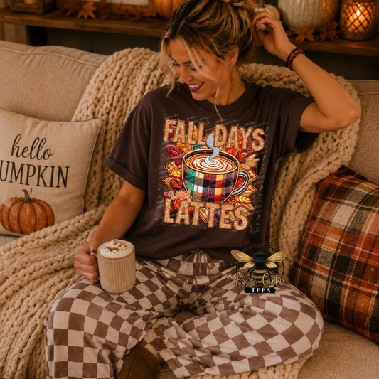 A woman sitting on a cozy couch wearing a dark brown graphic t-shirt that reads 'FALL DAYS LATTES' with a coffee cup design, paired with brown and beige checkered pajama pants. She is holding a mug of coffee, surrounded by autumn-themed pillows and a chunky knit blanket.