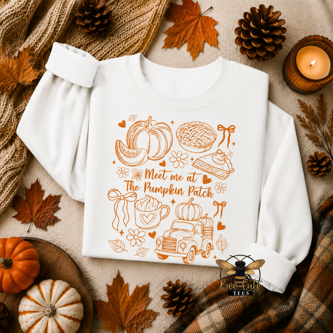 A white long-sleeve sweatshirt with a warm orange autumn-themed graphic reading 'Meet me at The Pumpkin Patch' and illustrated pumpkins, a truck, pie, and bows, styled in a flat lay with fall leaves, pinecones, a candle, and plaid fabric.