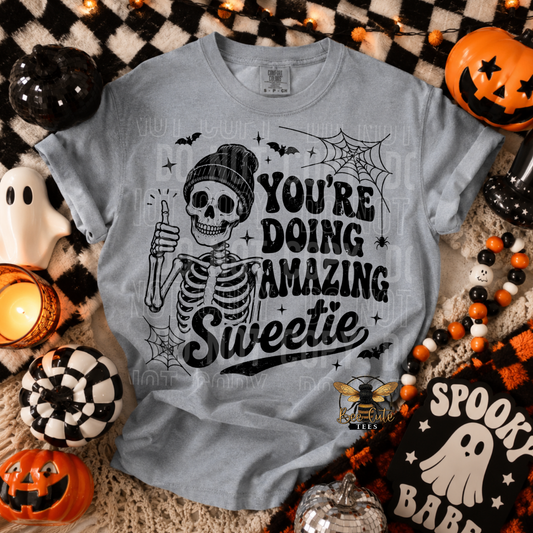 A grey crewneck t-shirt featuring a black graphic of a skeleton wearing a beanie and giving a thumbs-up, with the text 'YOU'RE DOING AMAZING Sweetie'. The shirt is displayed in a flat-lay Halloween setting with pumpkins, a ghost, and a checkered blanket.
