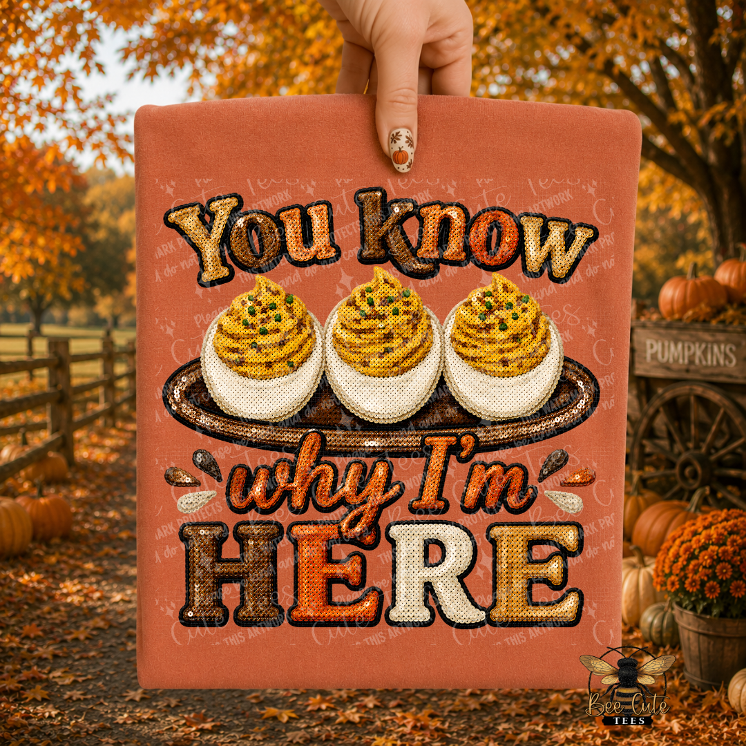 A hand holds up a folded orange t-shirt featuring the phrase 'You know why I'm HERE' with three deviled eggs on a platter. The background is a blurred autumn outdoor scene with pumpkins, fall leaves, and trees.