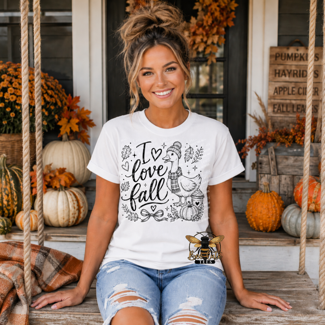 A woman sitting on a porch swing wearing a white graphic t-shirt with a black line art design that reads 'I love fall' with a turkey, pumpkins, and autumn leaves. The image has a digital watermark overlay of a bee on the bottom right.