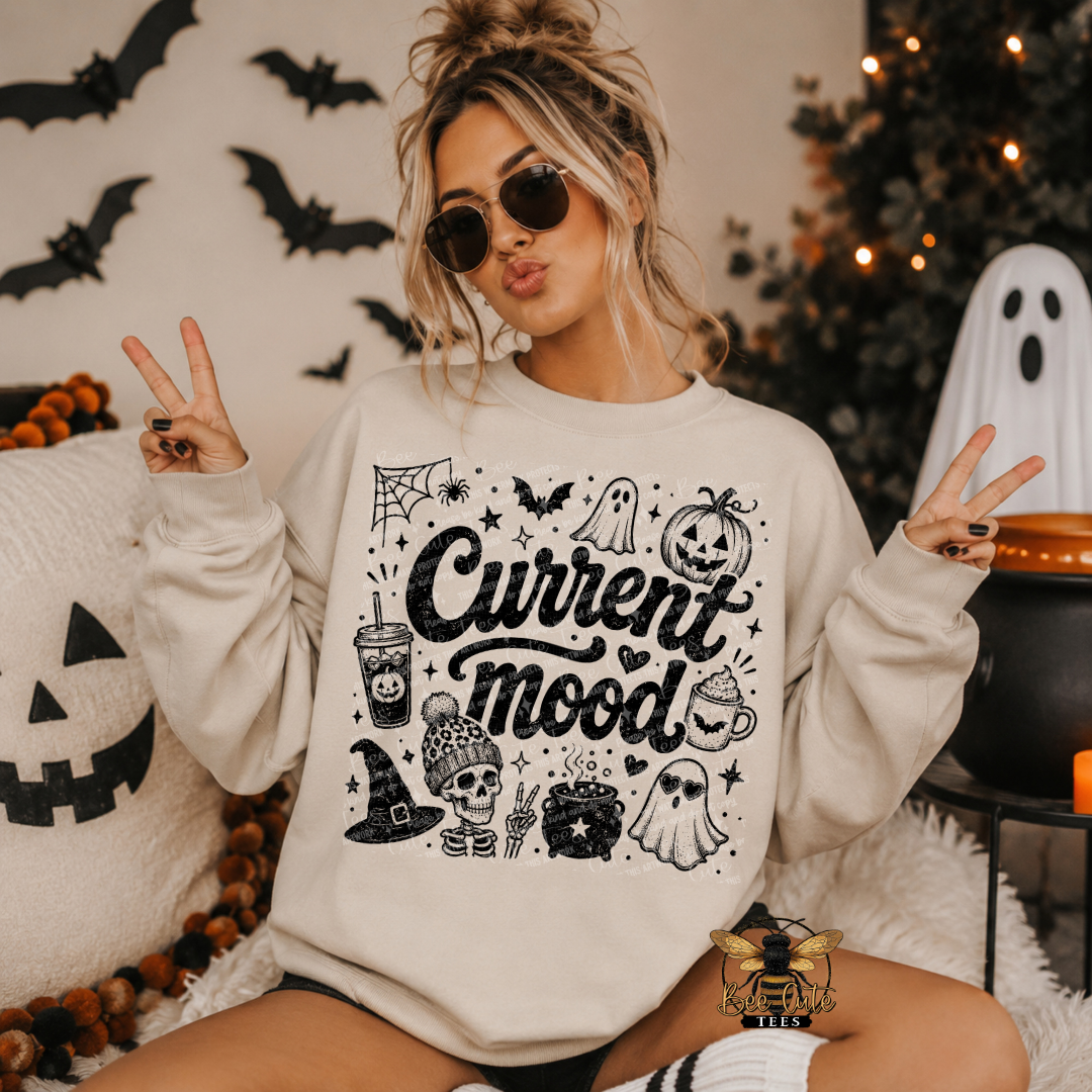 A woman wearing a cream-colored crewneck sweatshirt with a black 'Current mood' Halloween graphic, posing in a cozy, decorated Halloween room with jack-o'-lanterns, a ghost, and a Christmas tree in the background.