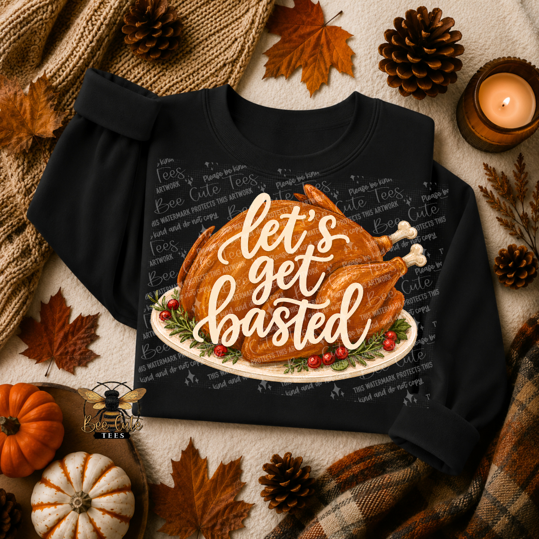 A black long-sleeve crewneck sweatshirt laid flat with a Thanksgiving-themed graphic of a roasted turkey on a platter and the phrase “let’s get baasted” in white script. The sweatshirt is surrounded by autumn props including pinecones, fall leaves, pumpkins, a candle, and a plaid blanket.