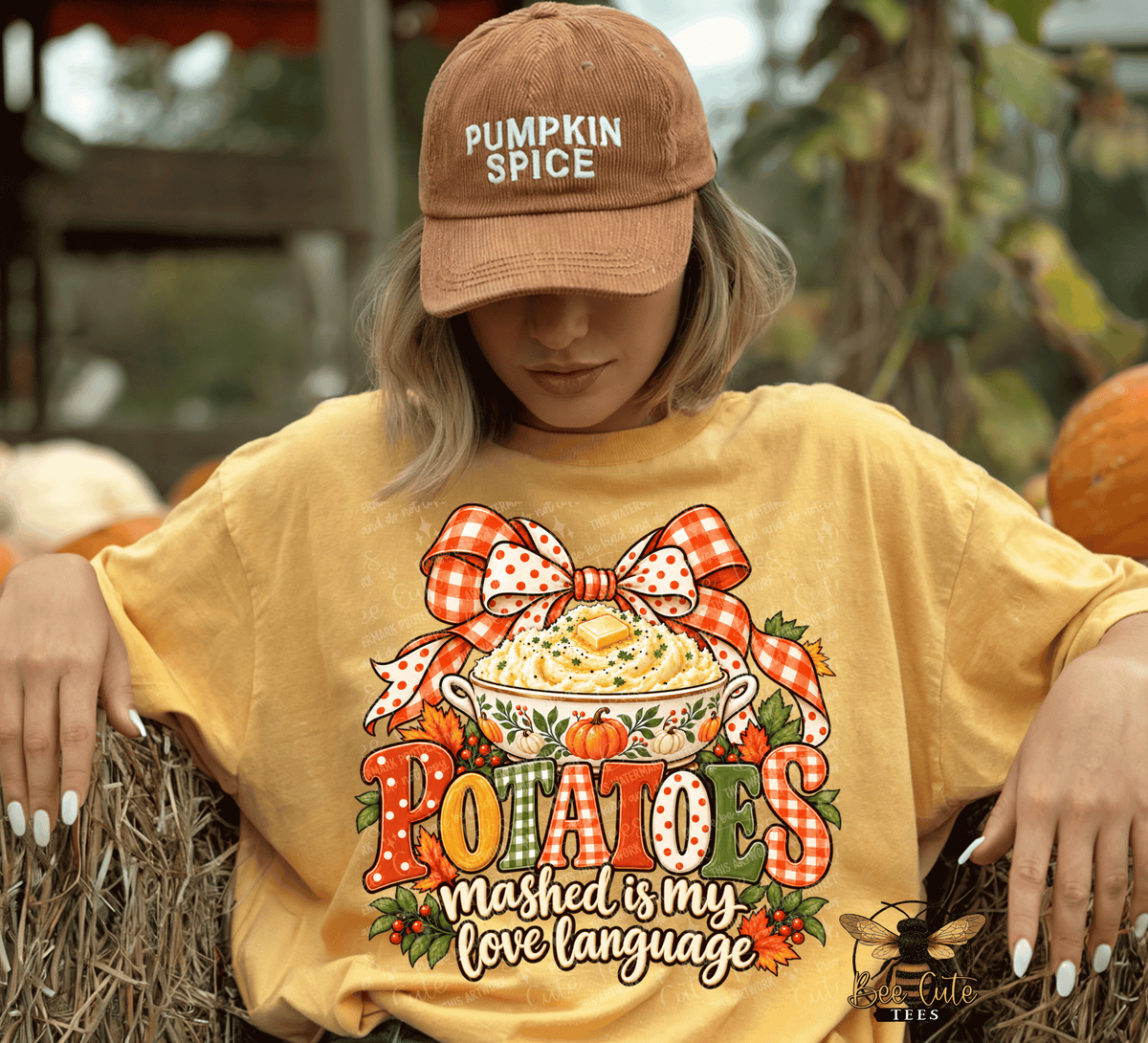 A woman wearing a brown 'Pumpkin Spice' baseball cap and a yellow graphic t-shirt with a fall-themed potato bowl design. She is sitting outdoors on a hay bale with pumpkins in the background.