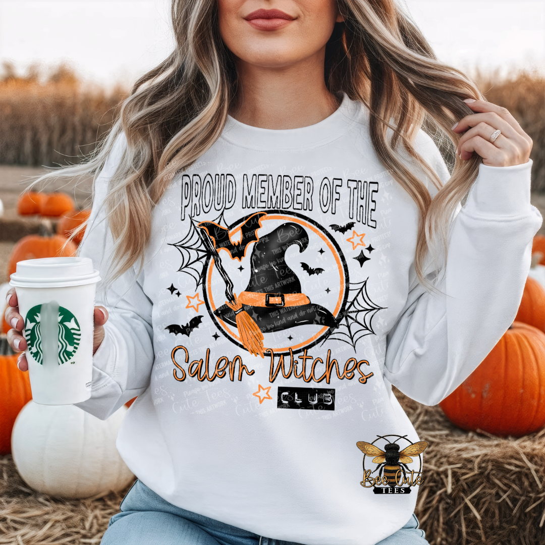 A woman in a white sweatshirt with a Halloween graphic reading 'PROUD MEMBER OF THE Salem Witches CLUB' stands in a pumpkin field holding a coffee cup. The sweatshirt features a witch hat, broom, bats, spiderwebs, and stars in orange, black, and gray.