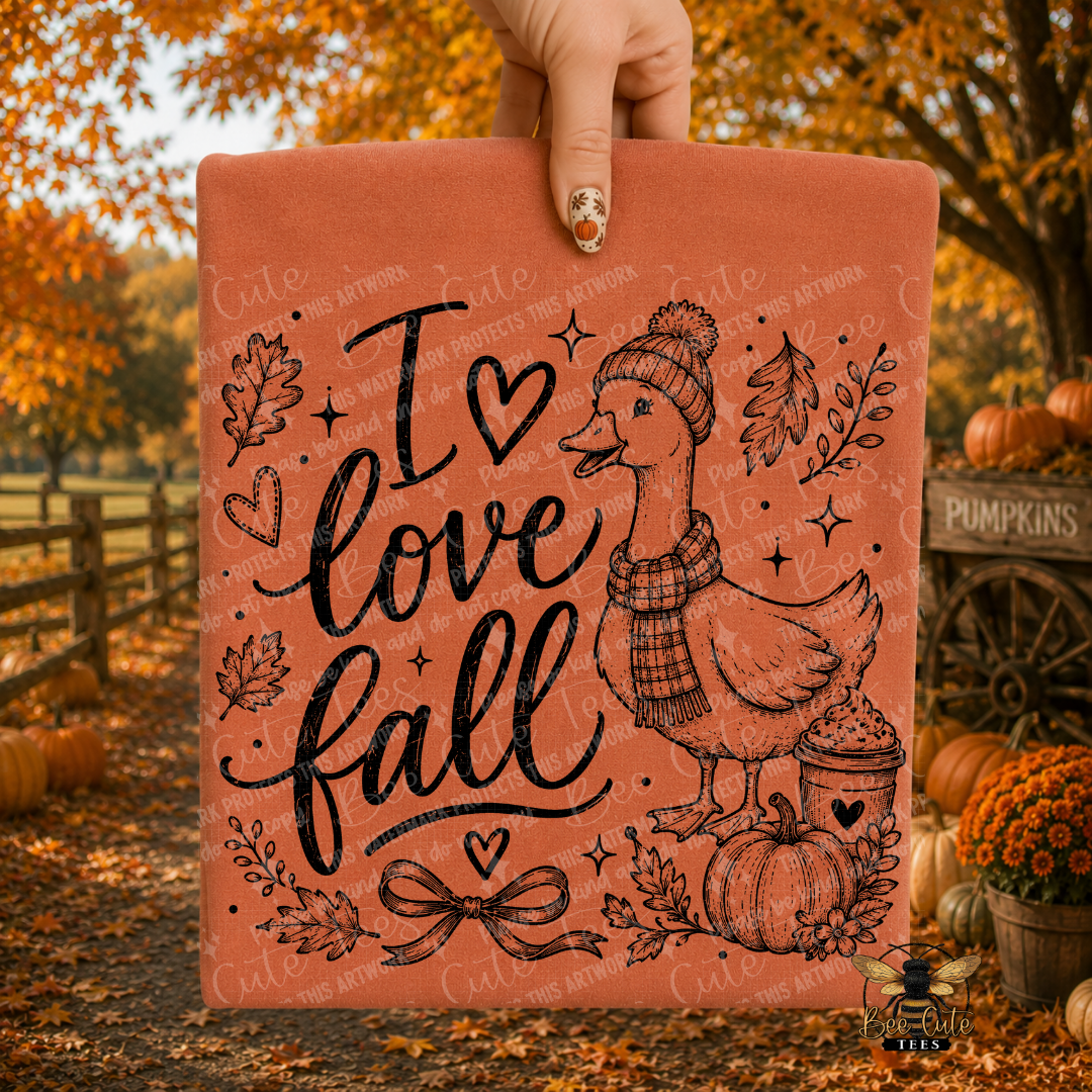 A hand holds a folded orange colored t-shirt featuring the phrase “I love fall” in black script, with a duck wearing a beanie and scarf, pumpkins, leaves, and small decorative motifs. The background shows an autumn outdoor scene with orange trees, fallen leaves, and pumpkins.