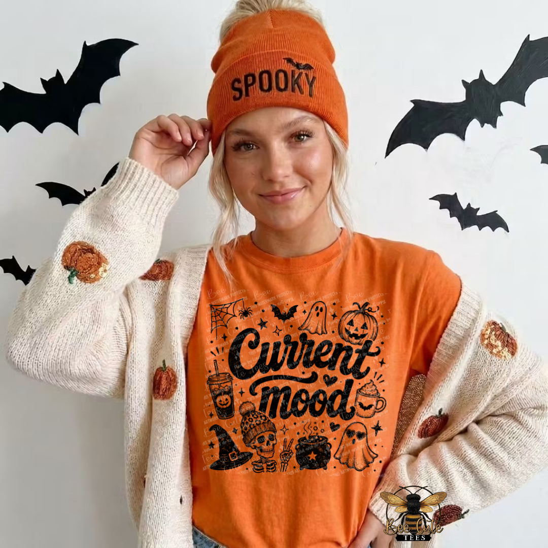 A woman wearing a bright orange beanie with 'SPOOKY' printed on it, an orange graphic sweatshirt that reads 'Current mood' with Halloween illustrations, and a cream knit cardigan with pumpkin patches, posed against a white wall decorated with black bat cutouts.