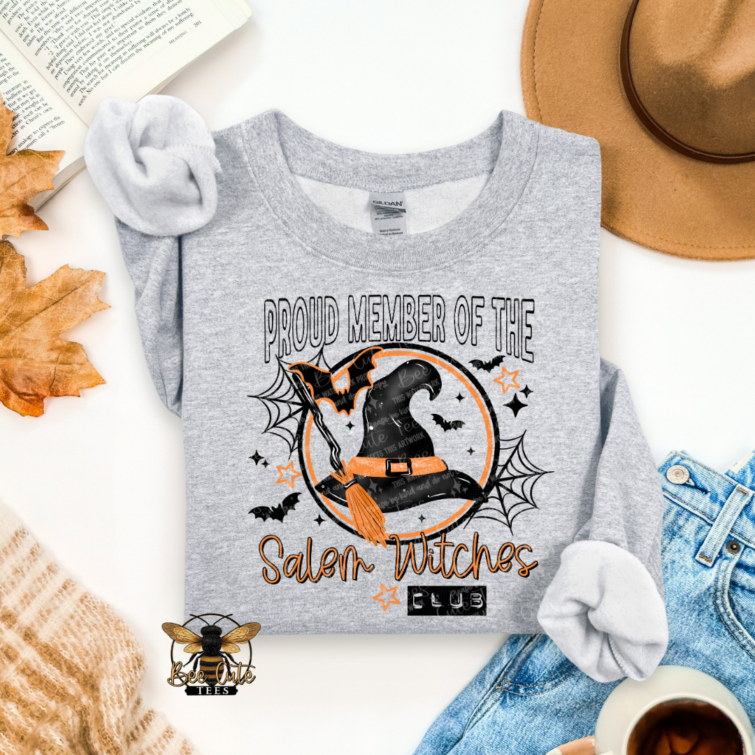 A heather gray long-sleeve t-shirt with a Halloween graphic reading 'PROUD MEMBER OF THE Salem Witches CLUB' featuring a witch hat, broom, bats, and spiderwebs, styled in a flat lay with jeans, a hat, leaves, and a cup.