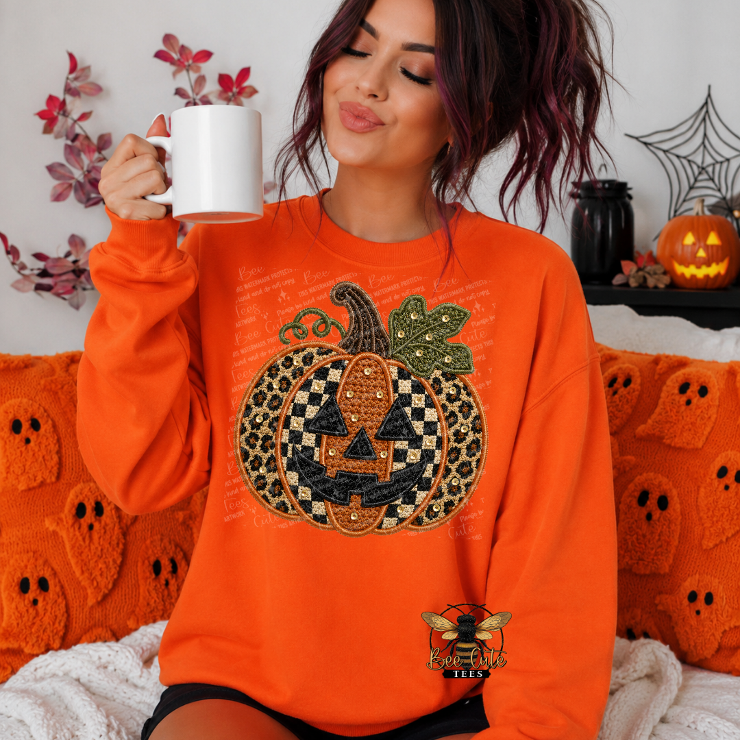 A woman wearing an orange sweatshirt with a large jack-o'-lantern graphic sits on a couch holding a white mug. The sweatshirt features a pumpkin design with leopard print, black and white checkers, and a bee patch near the bottom.