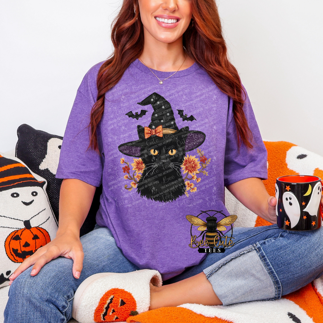 A woman wearing a purple t-shirt with a black cat wearing a witch hat graphic, sitting on a cozy white and orange couch with Halloween-themed pillows and holding a black mug with a white ghost design.