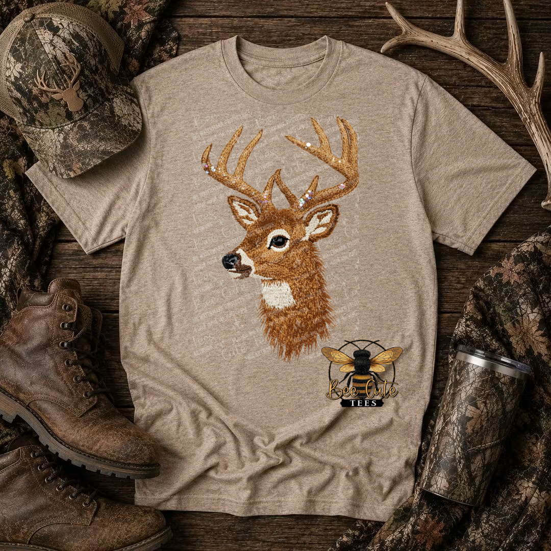A beige heather graphic t-shirt featuring a detailed embroidered deer head and a bee logo, styled in a rustic flat-lay with hunting boots, a camo cap, and antlers on a wooden background.