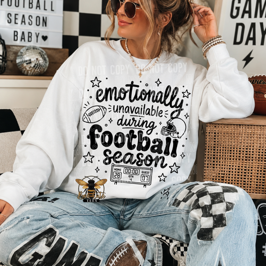 A woman wearing a white crewneck sweatshirt with a black graphic that reads 'emotionally unavailable during football season' with football-themed illustrations, paired with distressed light blue jeans and sunglasses.
