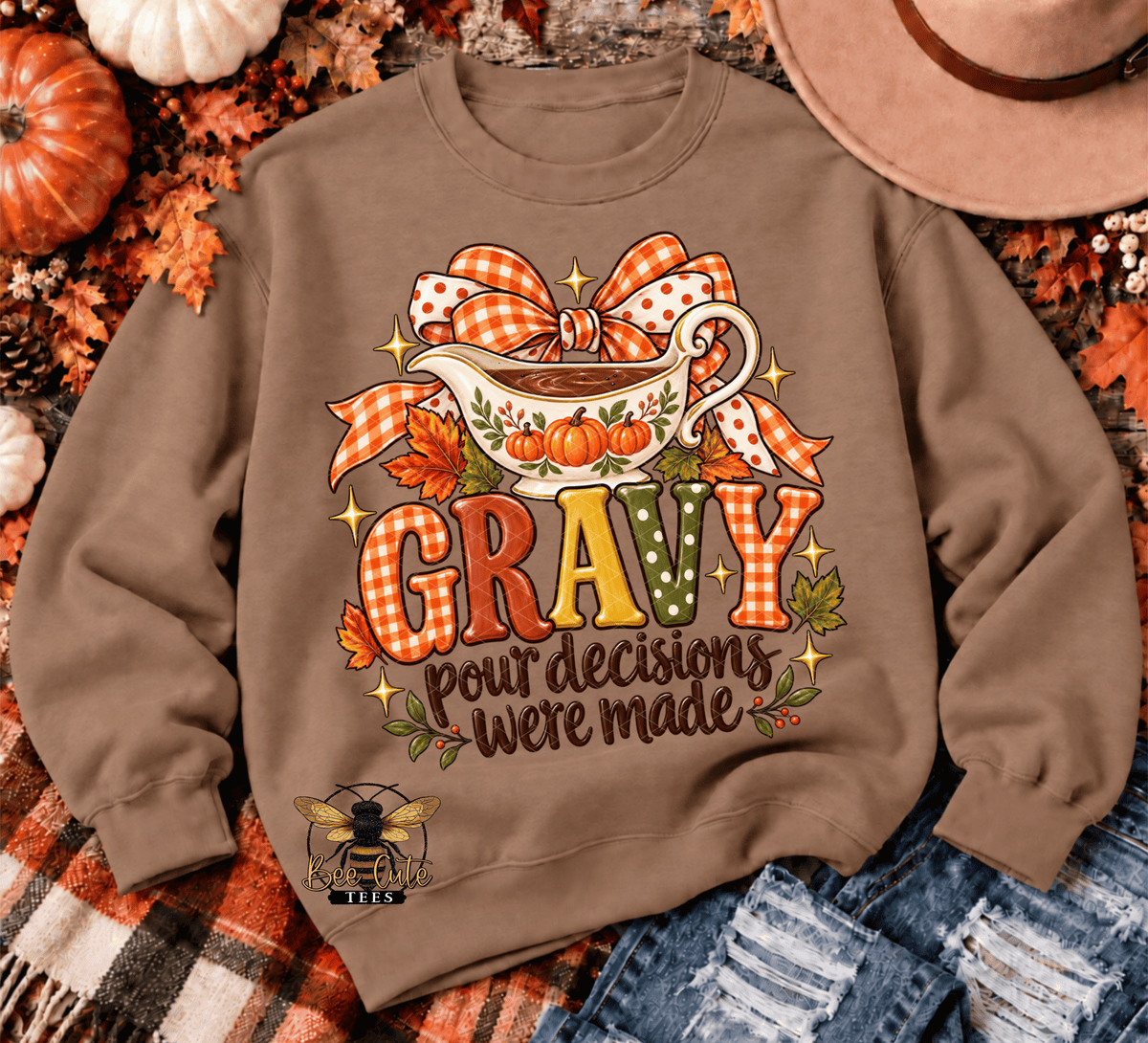 A brown crewneck sweatshirt with a fall-themed 'Gravy' graphic, laid flat on a plaid blanket with autumn props including pumpkins, leaves, a cowboy hat, and denim.