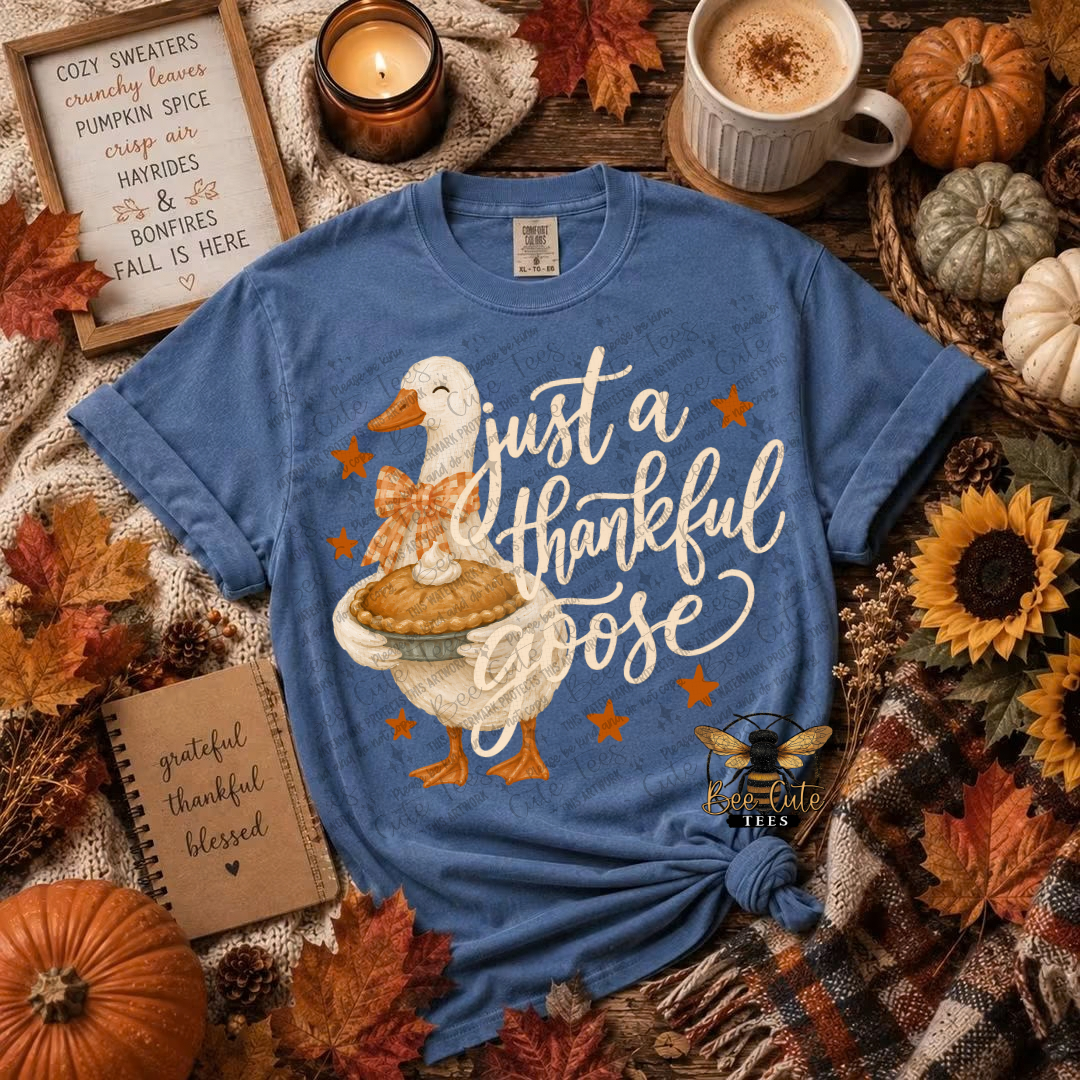 A blue graphic t-shirt featuring a cartoon Thanksgiving goose holding a pie with the text 'just a thankful goose' in white script, styled in a cozy autumn flat-lay with pumpkins, sunflowers, a candle, and a mug.