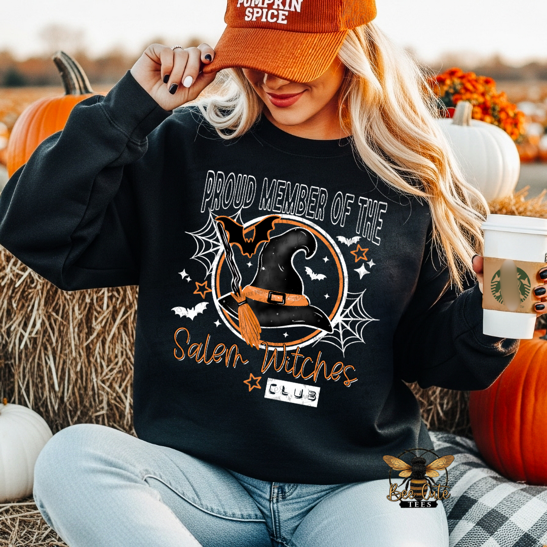 A woman sitting in a pumpkin patch wears a black crewneck sweatshirt with a Halloween graphic that reads 'PROUD MEMBER OF THE Salem Witches CLUB' and shows a witch hat, bats, and spiderwebs. She is wearing an orange 'Pumpkin Spice' cap and holding a  coffee cup.