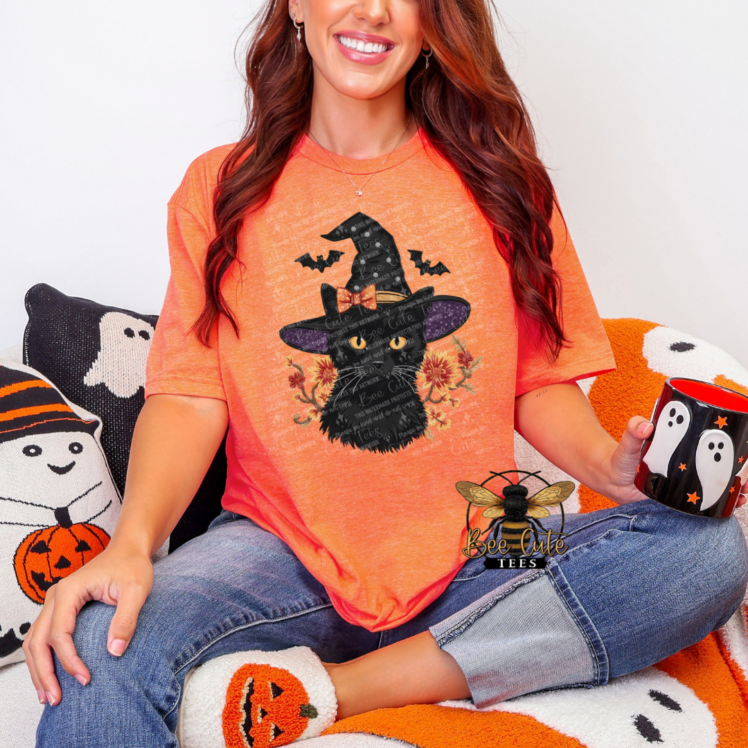 A woman wearing a bright orange t-shirt with a black cat wearing a witch hat graphic, sitting on a cozy white and orange couch with Halloween-themed pillows and holding a black mug with a white ghost design.