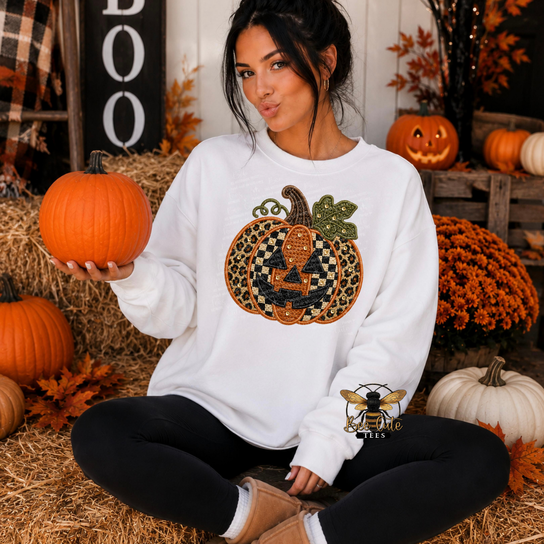 A woman sits cross-legged on a hay bale in a fall-themed setting, wearing a white sweatshirt with a large embroidered pumpkin design on the front. She holds a small orange pumpkin in one hand.