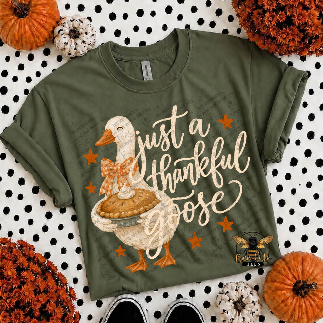 An olive green graphic t-shirt featuring a cartoon Thanksgiving goose holding a pie with the text 'just a thankful goose' in cream script and orange stars. The shirt is styled in a flat-lay with pumpkins, white and orange decorative gourds, and black-and-white sneakers on a black and white polka dot background.