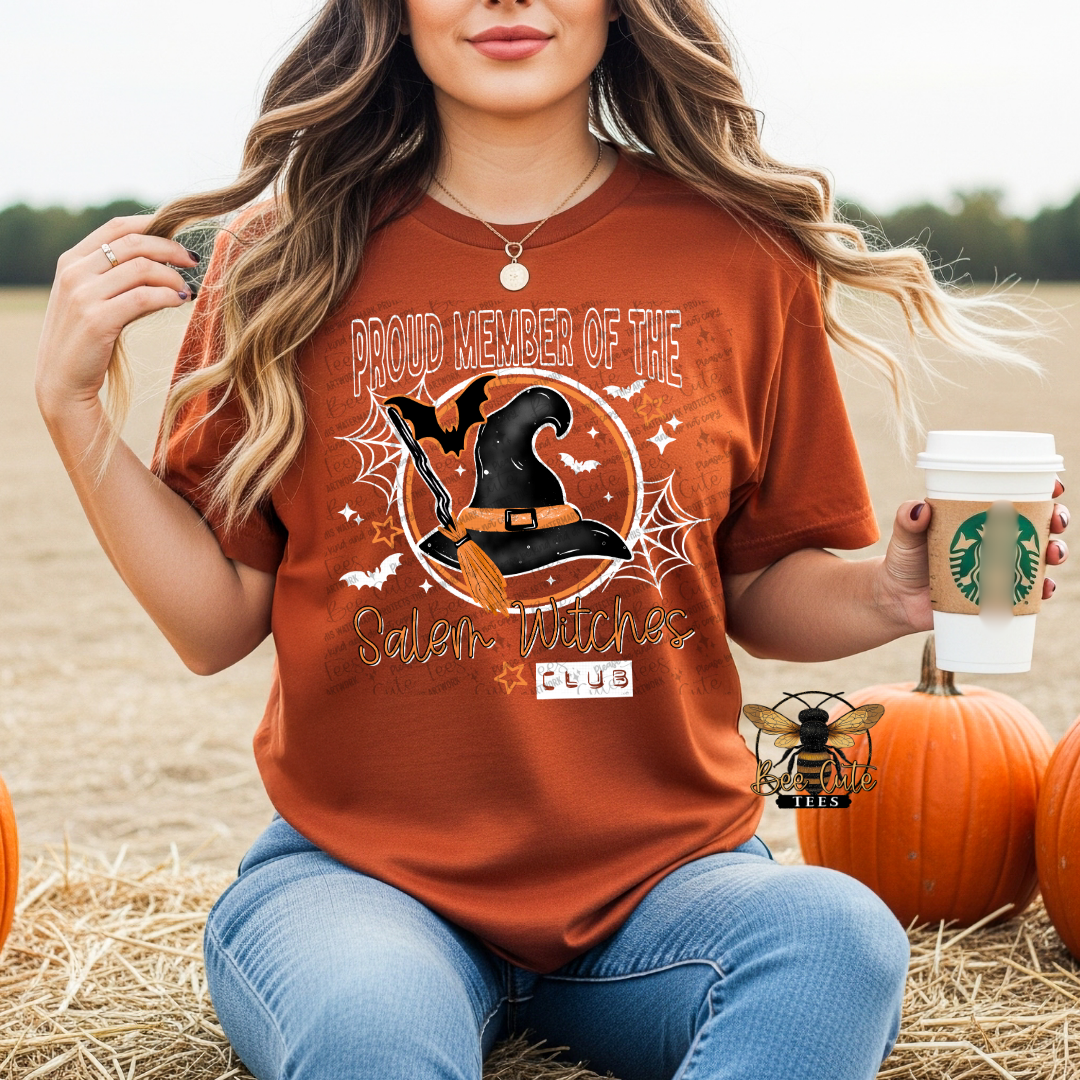 A woman wearing a rust-orange graphic t-shirt with a Halloween design that reads 'PROUD MEMBER OF THE Salem Witches CLUB' sits on a hay bale in a field. She is holding a coffee cup and surrounded by pumpkins.