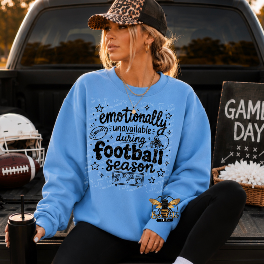 A woman wearing a light blue crewneck sweatshirt with a black football-themed graphic sits in the bed of a truck. The sweatshirt features the text 'emotionally unavailable during football season' with illustrations of a football, a helmet, and a scoreboard.