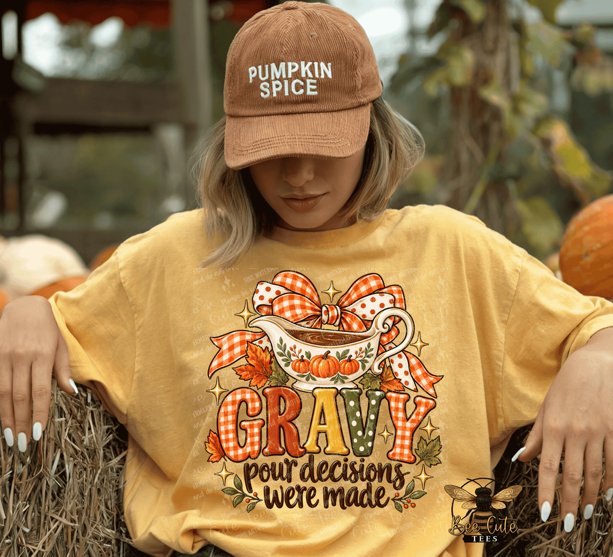 A woman wearing a brown 'Pumpkin Spice' baseball cap and a yellow graphic t-shirt sitting on a hay bale outdoors. The t-shirt features a fall-themed graphic with a coffee cup, pumpkins, gingham ribbon, and the text 'GRAVY pour decisions were made'.