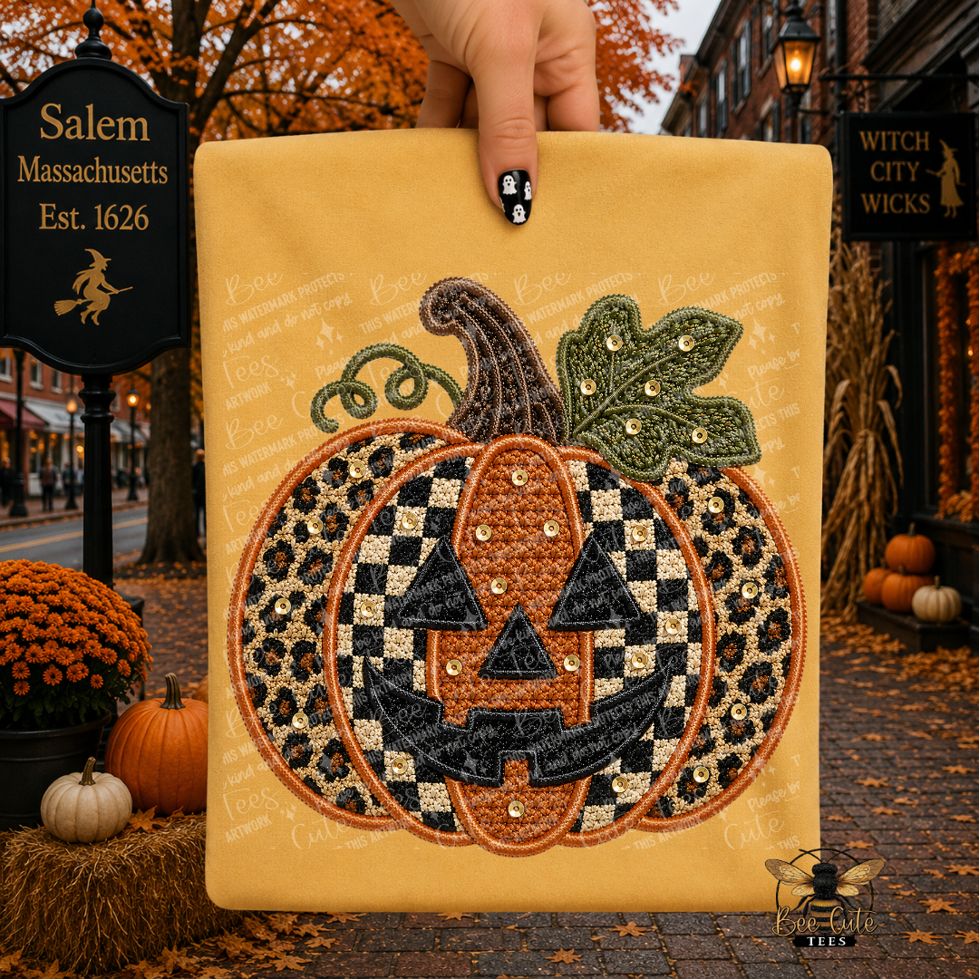 A hand holds a mustard yellow folded t-shirt featuring a large embroidered jack-o'-lantern design with black-and-white checkered and leopard-print sections, a green leaf, and small rhinestone accents. The background is a styled autumn street scene with fall leaves, pumpkins, and storefront signs.