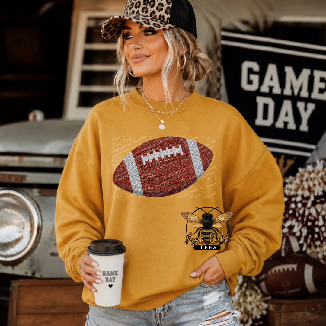A woman wearing a mustard yellow crewneck sweatshirt with a football graphic on the chest and a bee logo, holding a coffee cup in a rustic, sports-themed setting.