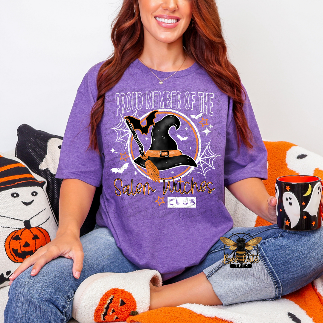 A woman wearing a purple graphic t-shirt with a Halloween design that reads 'PROUD MEMBER OF THE Salem Witches CLUB'. She is sitting on a cozy blanket with Halloween-themed pillows and slippers, holding a black mug with a ghost design.