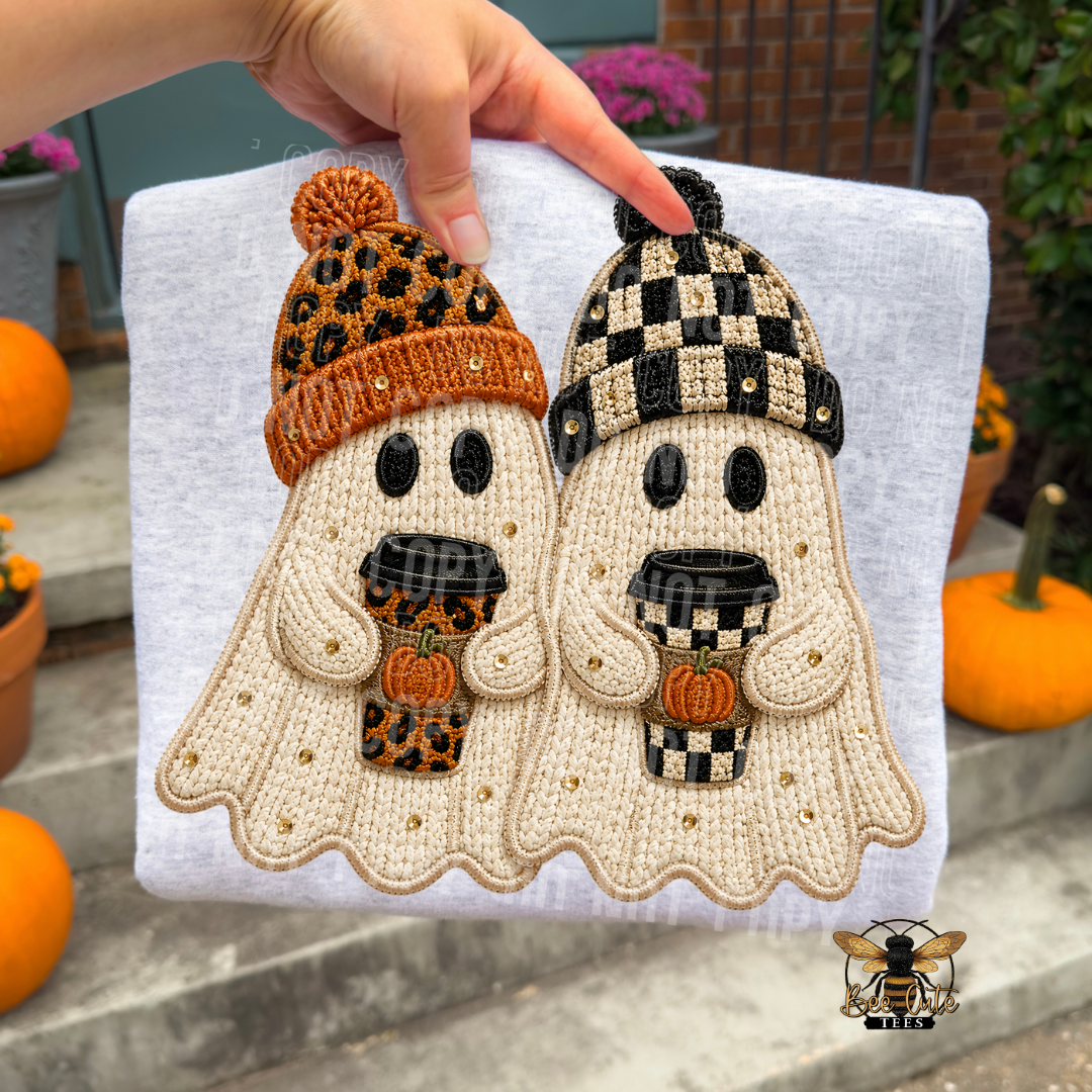 A hand holds a white t-shirt featuring a crochet-style graphic of two cute ghost characters wearing cozy autumn hats and holding coffee cups with pumpkins. The background shows outdoor patio elements like pumpkins and plants.