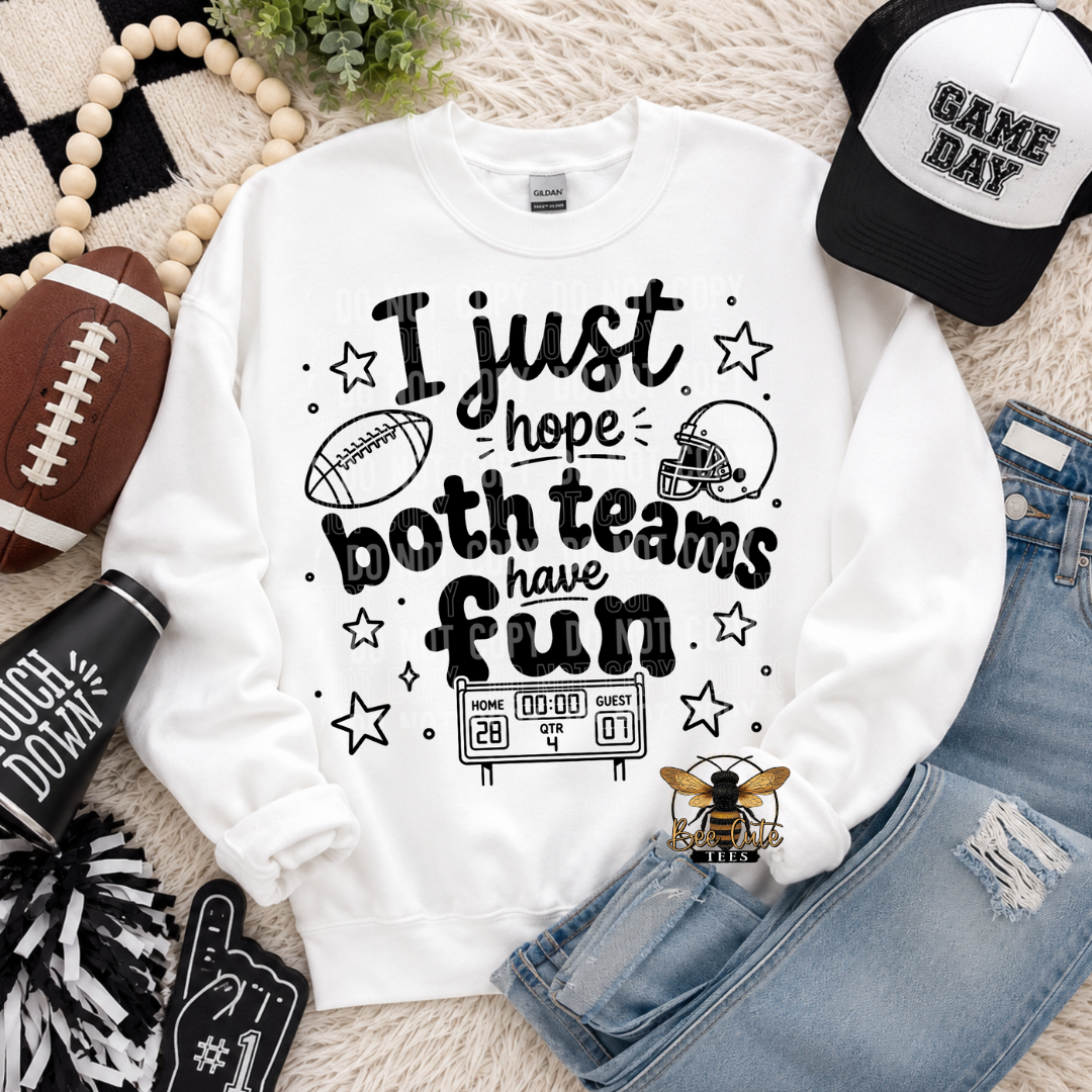 A white crewneck sweatshirt with a black graphic text reading 'I just hope both teams have fun' surrounded by football-themed doodles, stars, and a scoreboard. The sweatshirt is styled in a flat lay with a black and white 'GAME DAY' baseball cap, distressed blue jeans, a football, a pom-pom, a black foam finger, and a small bee logo on a beige textured rug.