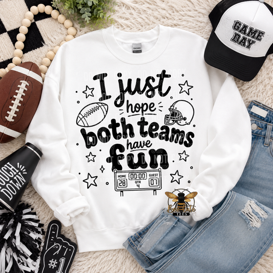 A white crewneck sweatshirt with a black graphic text reading 'I just hope both teams have fun' surrounded by football-themed doodles, stars, and a scoreboard. The sweatshirt is styled in a flat lay with a black and white 'GAME DAY' baseball cap, distressed blue jeans, a football, a pom-pom, a black foam finger, and a small bee logo on a beige textured rug.