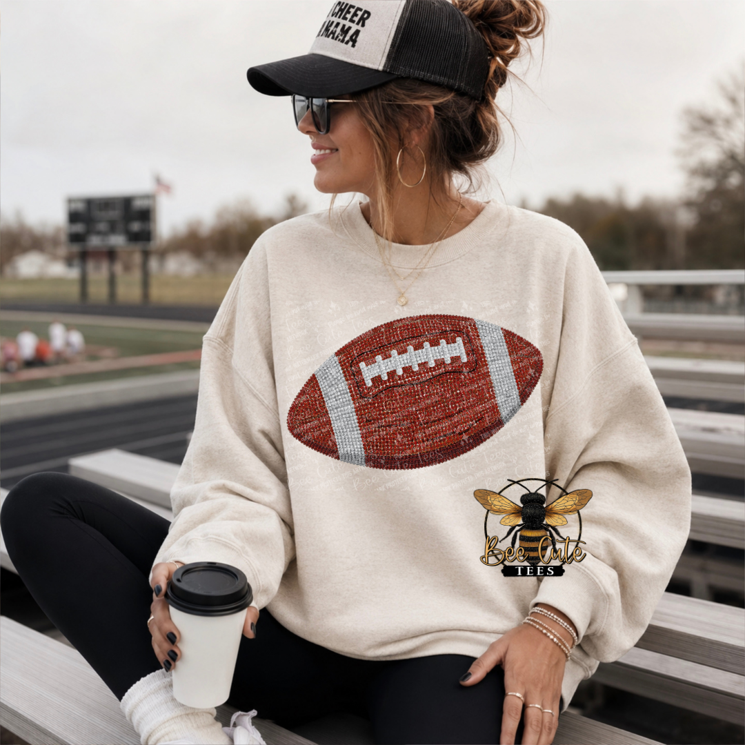 A woman sitting on outdoor bleachers wearing a cream sweatshirt with a large football graphic on the front, black leggings, a black cap, and holding a coffee cup.