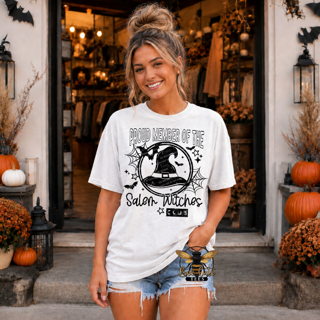 A woman stands in front of a storefront doorway wearing a white graphic T-shirt with a black witch-themed print reading 'Proud Member of the Salem Witches Club' and denim shorts. Pumpkins, fall foliage, lanterns, and a shop entrance are visible in the background.