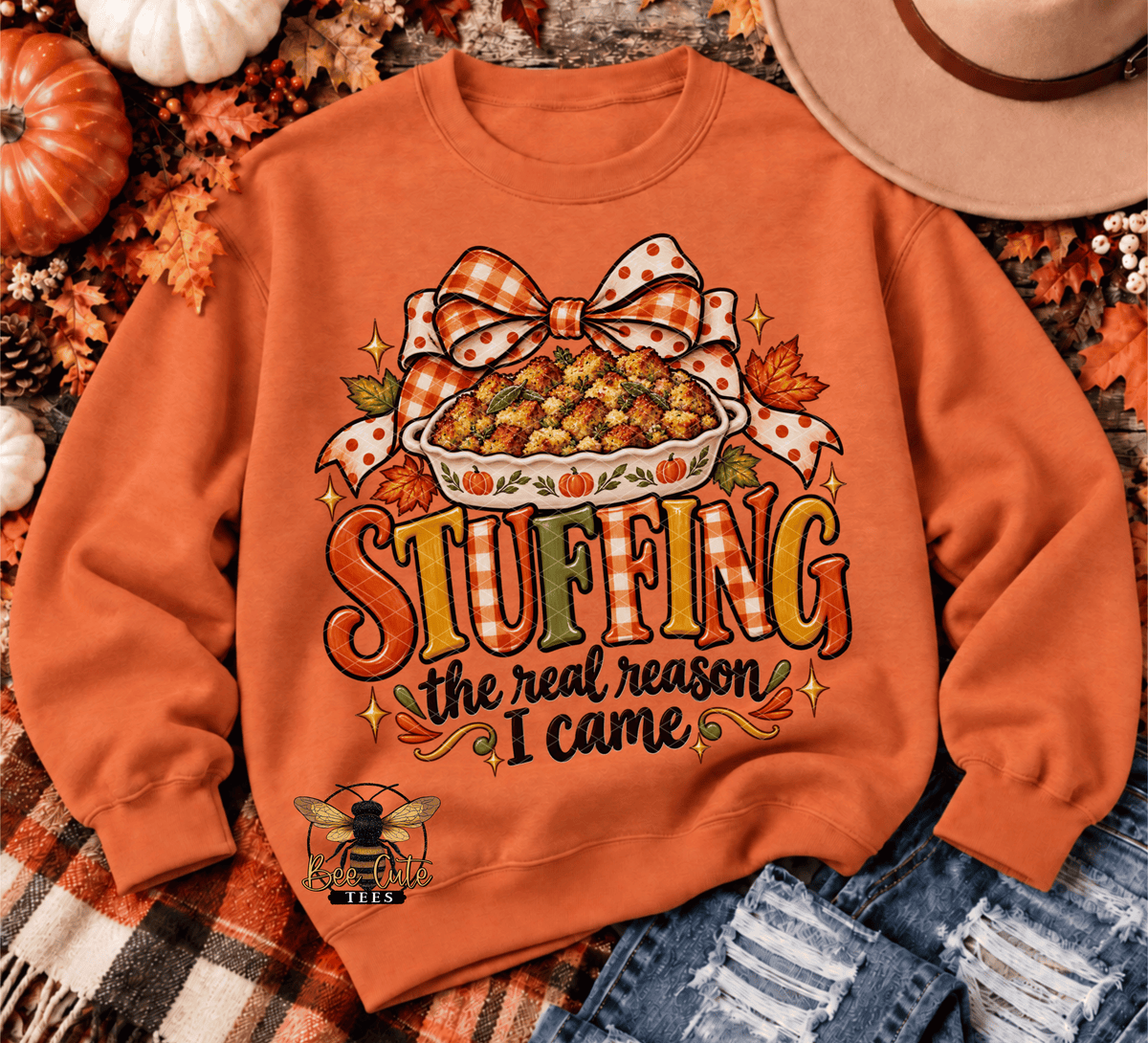 An orange crewneck sweatshirt with a Thanksgiving-themed graphic reading 'STUFFING the real reason I came' beneath a pie illustration and bow. The sweatshirt is styled in a flat lay with autumn leaves, pumpkins, a plaid blanket, denim, and a hat.