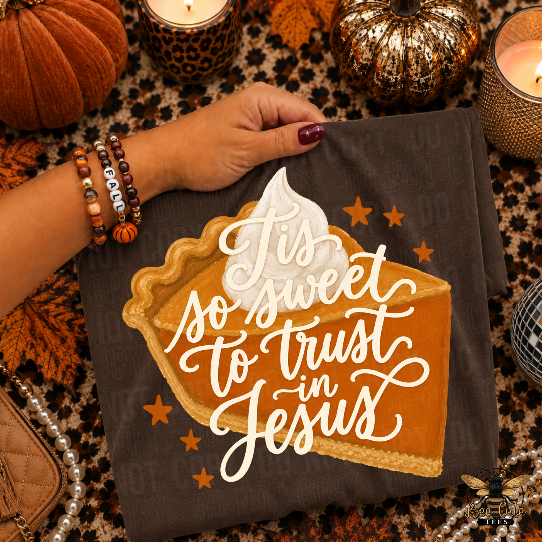 A folded brown T-shirt is displayed on a patterned surface with seasonal props. A hand with purple nail polish holds the top edge.