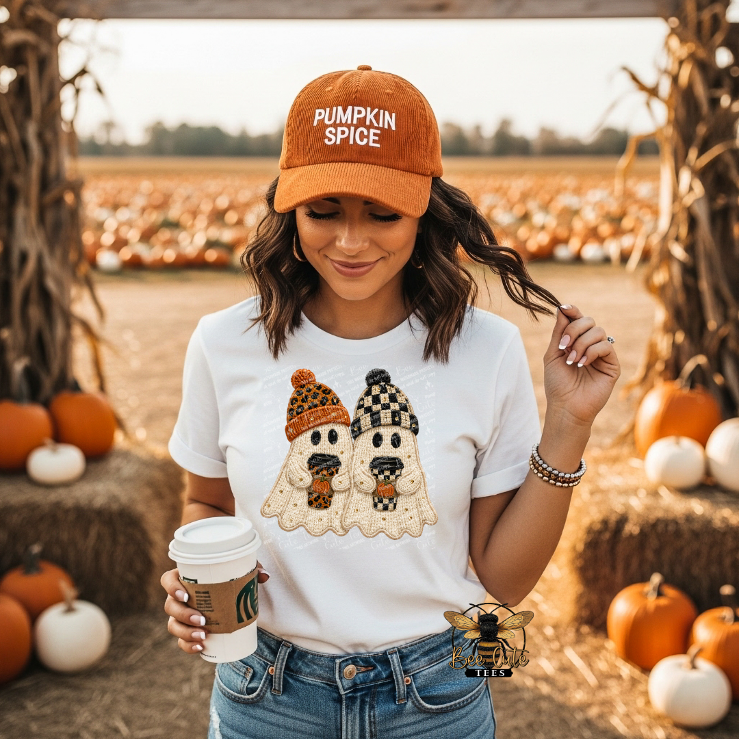 A woman standing in a pumpkin patch wearing an orange 'Pumpkin Spice' baseball cap and a white graphic t-shirt featuring two ghost characters. She is holding a coffee cup.