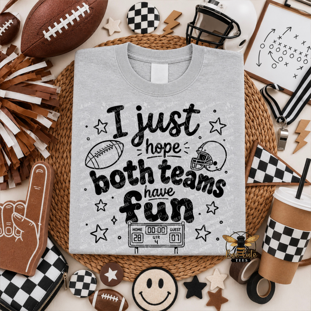 A light gray graphic t-shirt featuring a black football-themed design with the text 'I just hope both teams have fun' and illustrations of a football, helmet, stars, and scoreboard. The shirt is presented in a styled flat-lay arrangement with various football accessories, a foam finger, and checkered items on a neutral background.