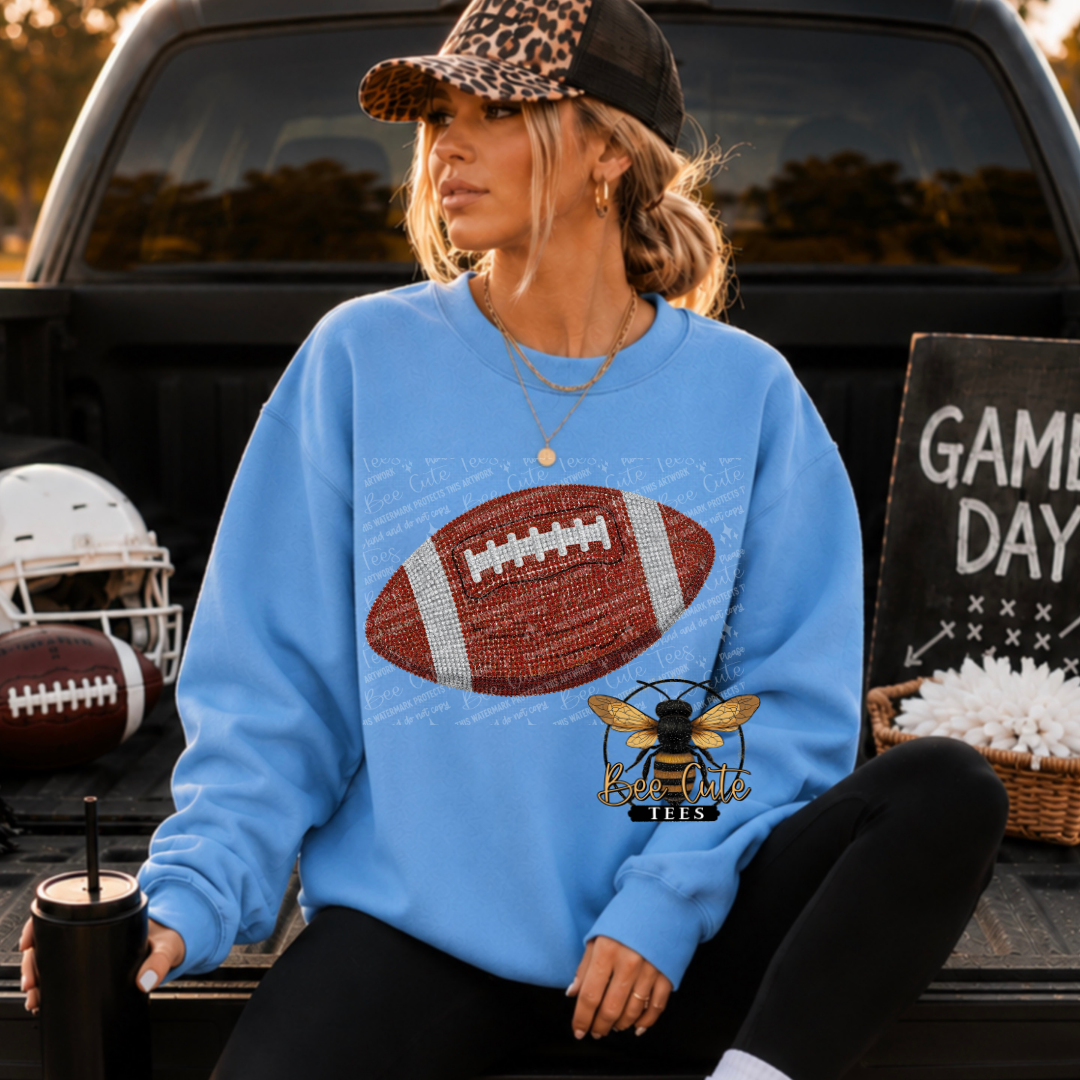 A woman sitting in the bed of a pickup truck wearing a light blue crewneck sweatshirt with a large football graphic and a bee logo . A football helmet, a football, and a 'Game Day' sign are visible around her.