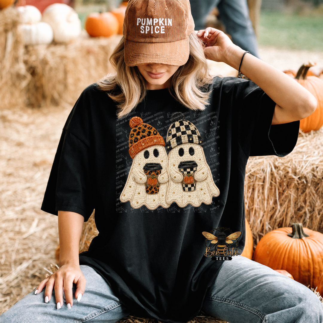 A woman sitting outdoors on hay sits wearing a black graphic t-shirt with a cartoon ghost design, a brown 'Pumpkin Spice' baseball cap, and blue jeans. Pumpkins and hay bales are visible in the background.