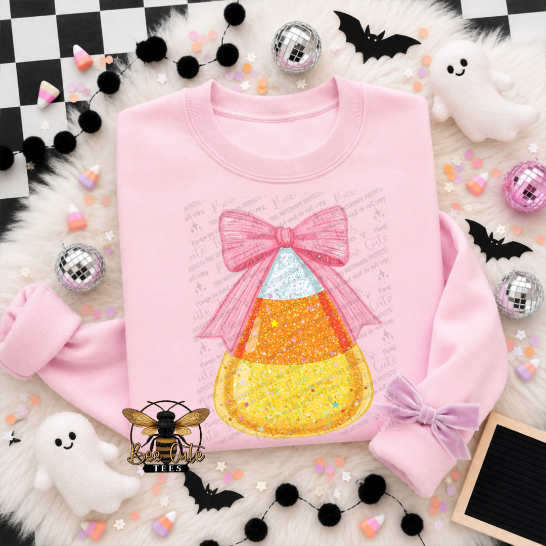 A pink long-sleeve sweatshirt laid flat on a white faux-fur surface, featuring a large candy corn graphic with a pink bow and glittery yellow, orange, and white sections. The sweatshirt is styled with Halloween props including black pom-pom garlands, silver disco balls, white ghost cutouts, black bat silhouettes, pastel candy pieces, and a checkered corner backdrop.