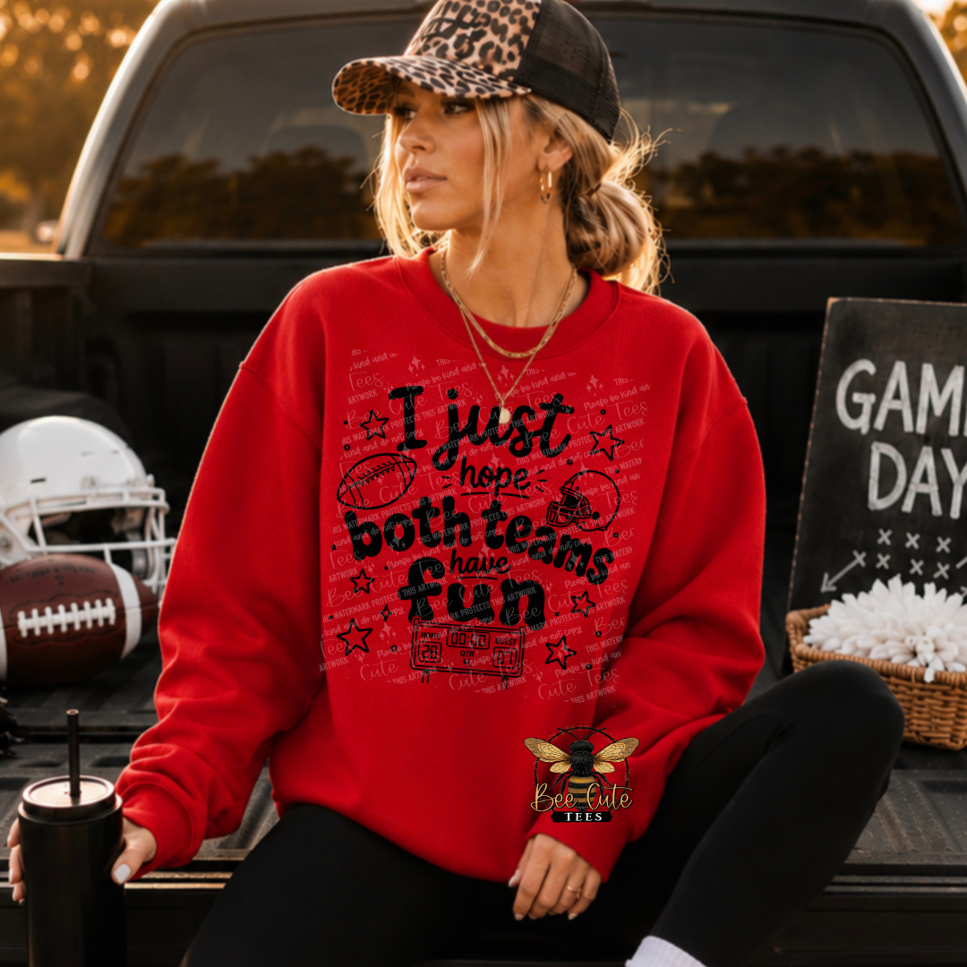 A woman wearing a red graphic sweatshirt with a football theme sits in the bed of a truck. The sweatshirt features a black graphic that reads 'I just hope both teams have fun' surrounded by football illustrations.