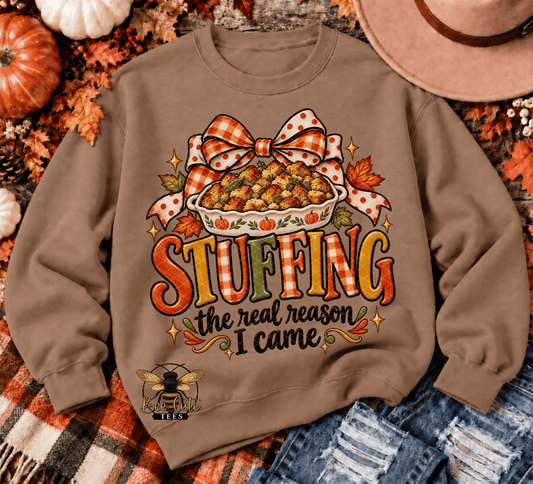 A brown crewneck sweatshirt with a colorful autumn-themed graphic reading 'STUFFING the real reason I came' with a pie, bow, and fall leaves. The sweatshirt is styled in a flat-lay with a plaid blanket, jeans, a hat, and pumpkins.