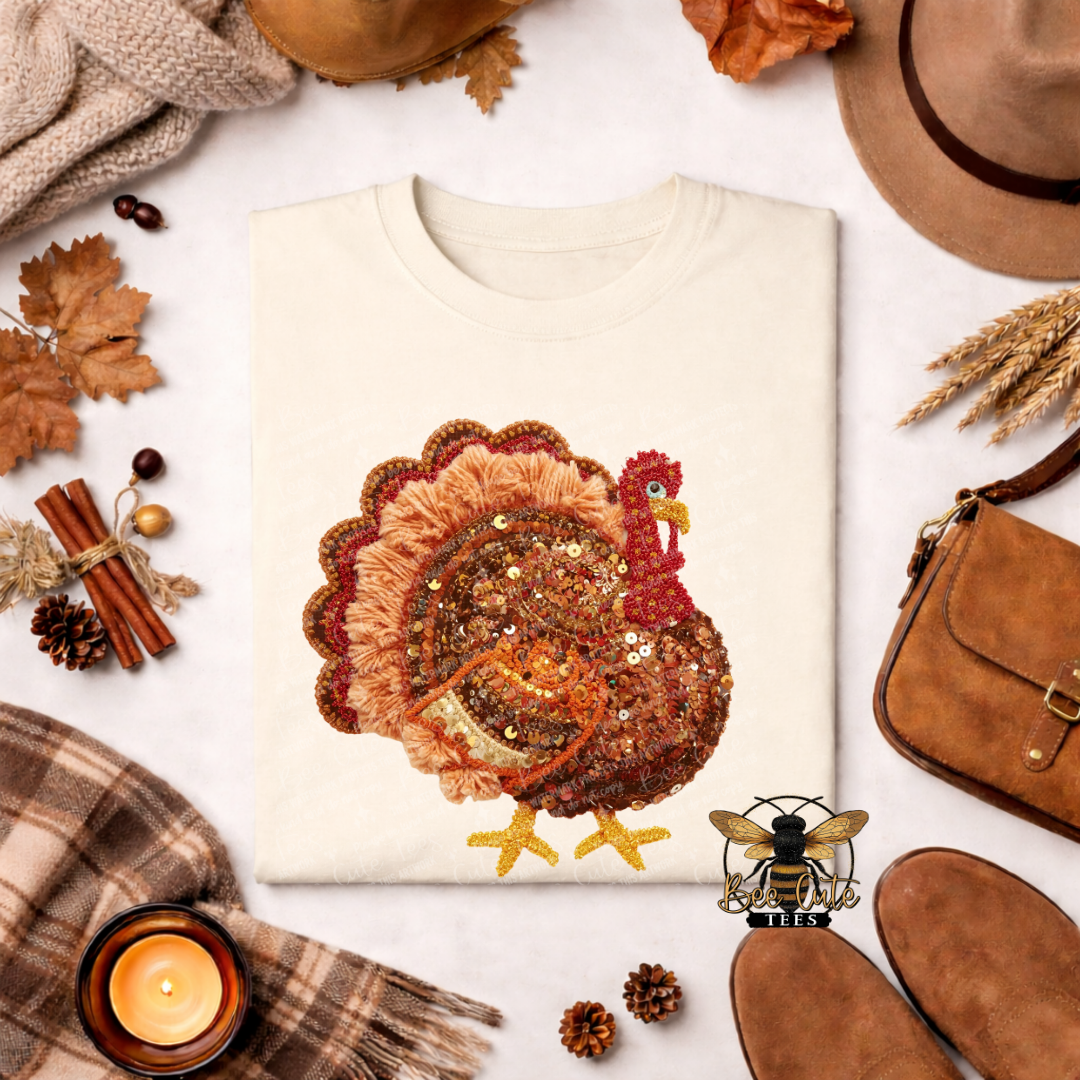 A folded cream-colored t-shirt with a large, detailed turkey design made from brown sequins and beads, surrounded by autumn-themed props like a plaid scarf, a candle, pinecones, and a brown suede bag on a white background.